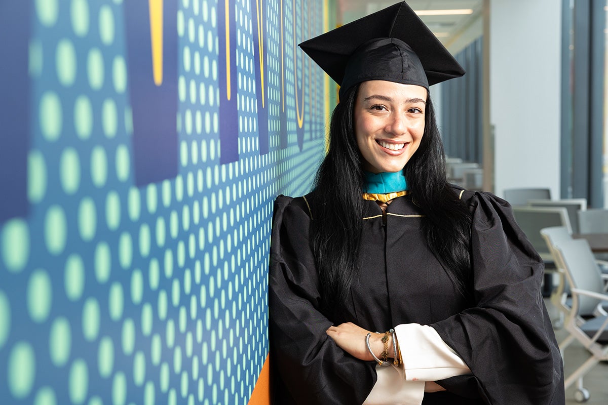 Woman wearing black cap and gown smiles as she leans next to blue polkadot wall