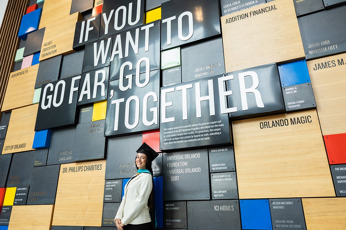 Woman in white blazer and traditional black grad cap stands in front of mural that reads "If you want to go far, go together."