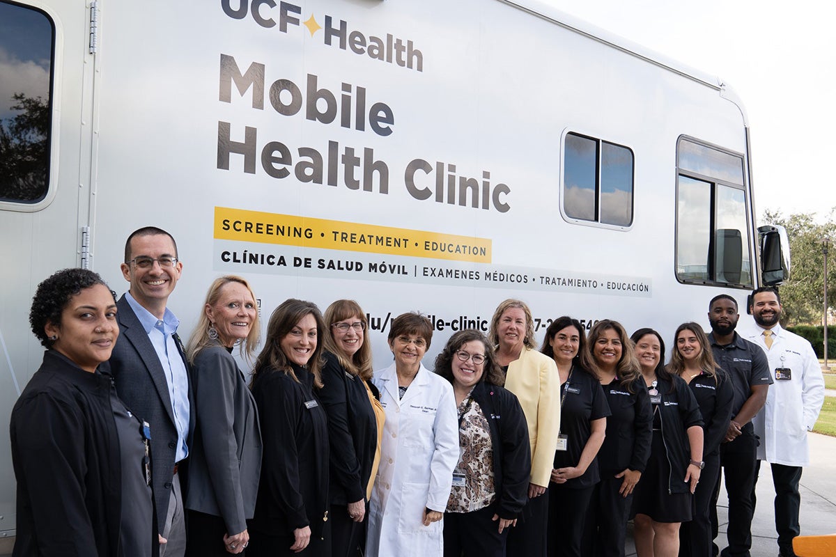 A dozen adults stand in front of white vehicle with mobile health clinic branding 