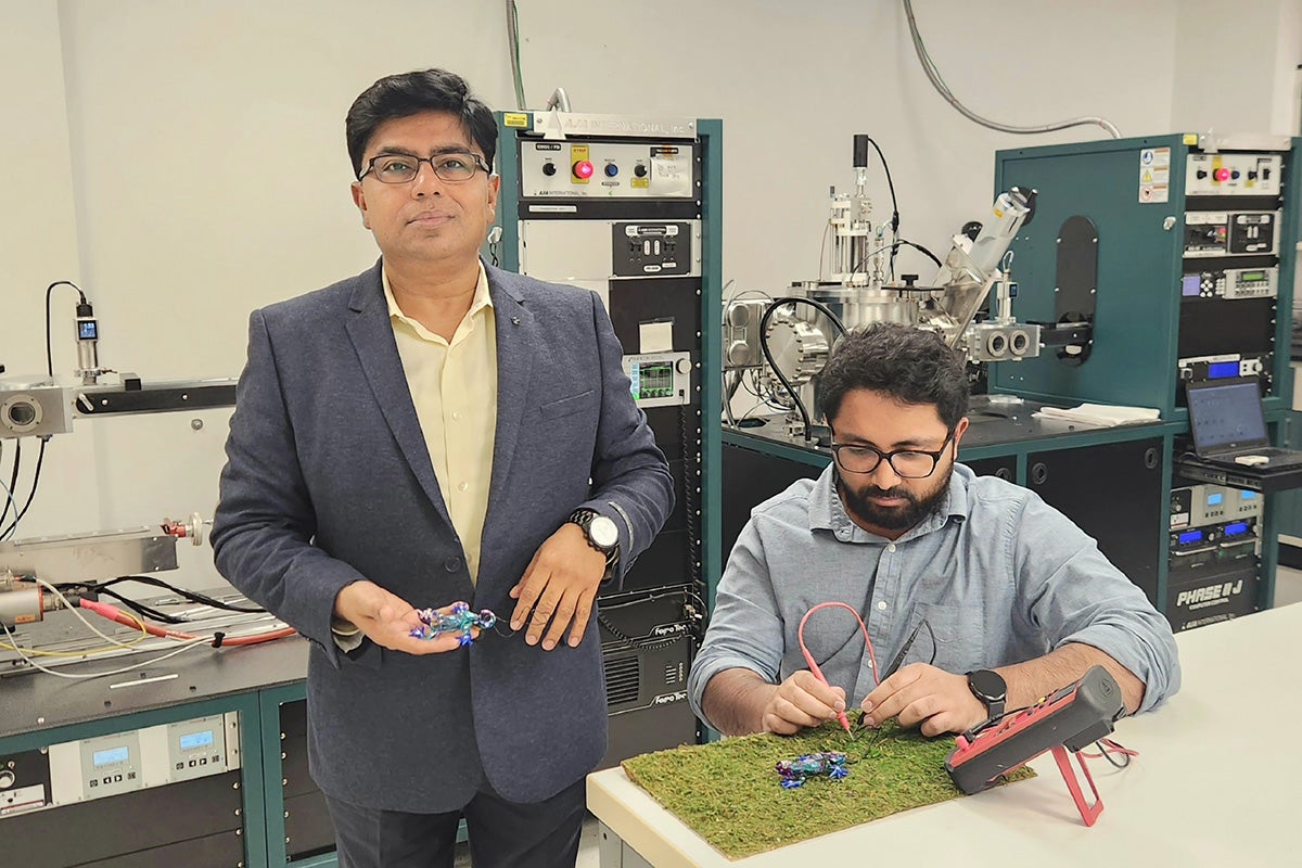 Debashis Chanda in a gray suit holds a colorful small gadget in his hand while standing in a lab next to a table and bench, where a bearded man works on a similar gadget