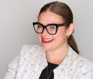 Brunette woman with her hair in a ponytail, black glasses and a white business coat smiles