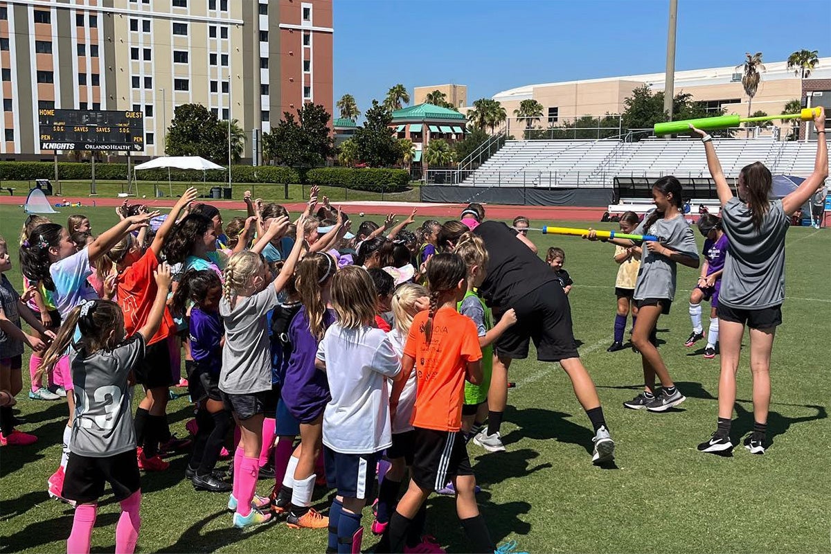 Young children stand on soccer field with camp counselors squirting water from pool noodle squirters on sunny day