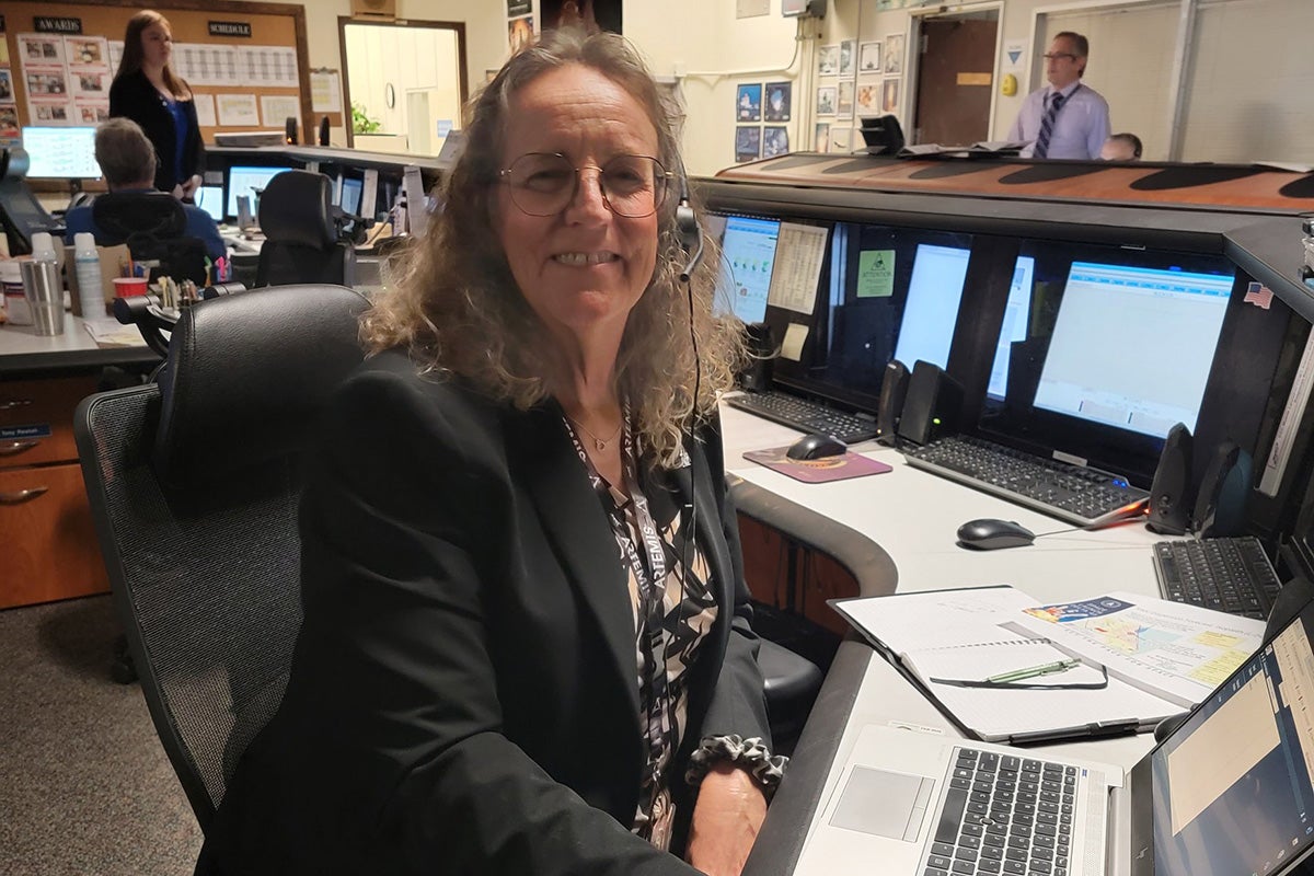 Woman with curly, sandy blonde hair in business jacket sits at console with screens and keyboard