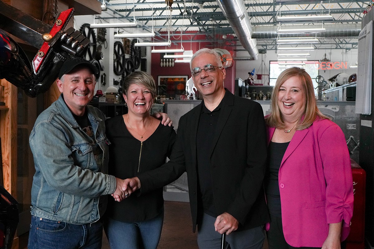 A portrait of two men and two women standing in warehouse with technical equipment behind them as the two men shake hands.