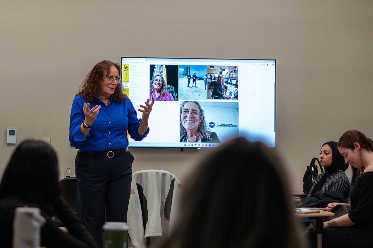 Woman in blue button down long sleeve shirt and blank pants stands in front of screen at front of classroom, speaking to women seated