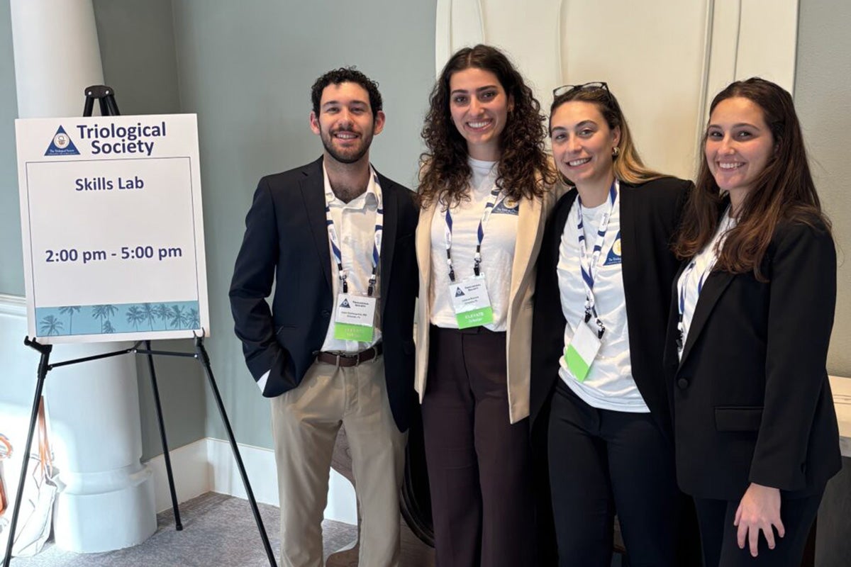Three women and one man all in blazers stand next to easel displaying sign that reads Triological Society skills lab 2-5 p.m.