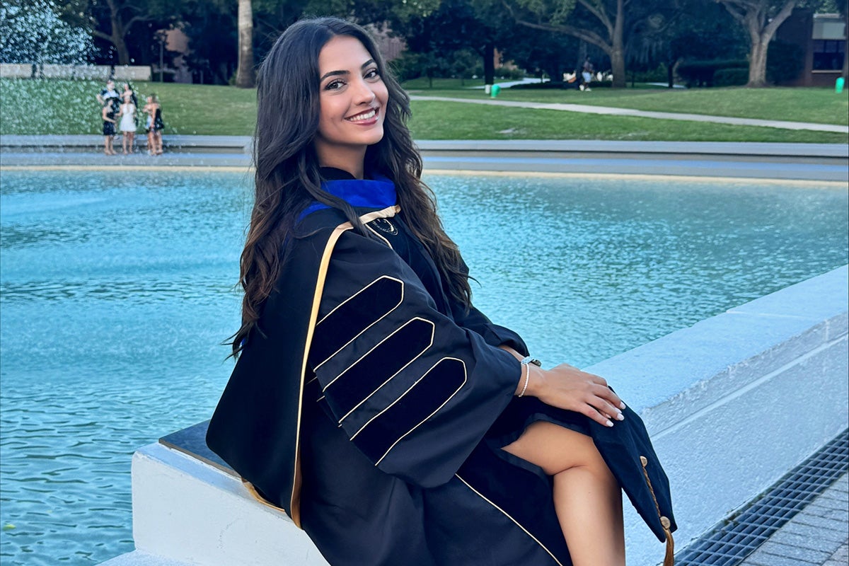 UCF alumna Ilkin Isler, in graduation regalia, sits on a stone bench near UCF’s Reflecting Pond.