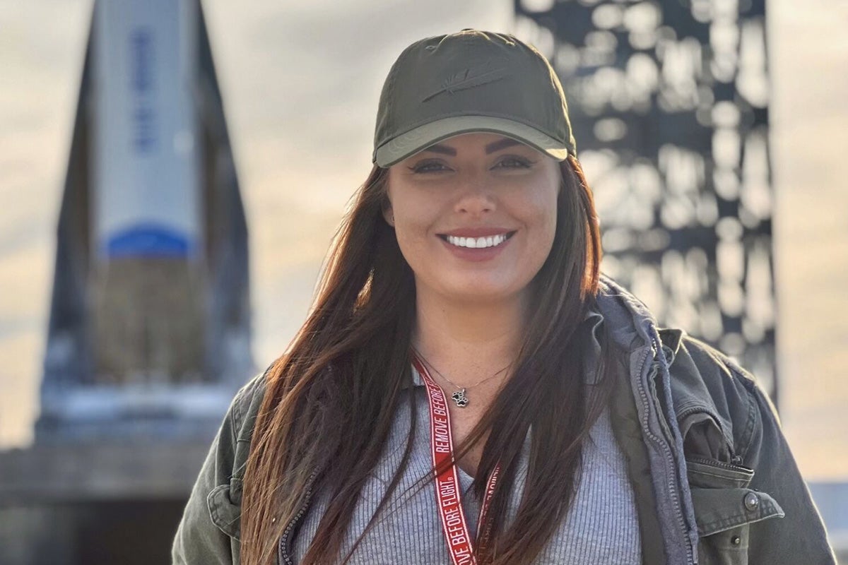 Woman wearing olive green baseball cap smiles as she stands in front of New Glenn rocket