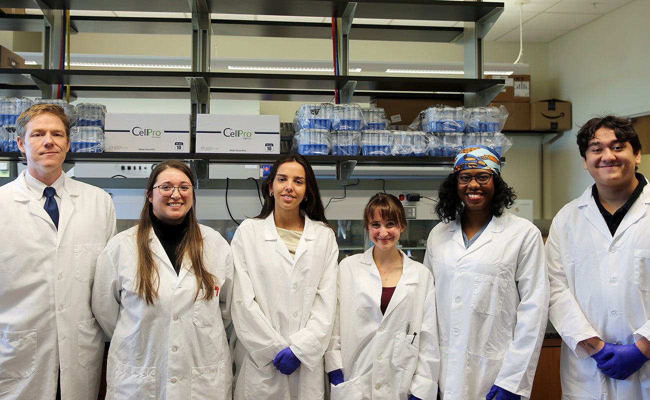 Six individuals in lab coats pose shoulder to shoulder in lab with shelving behind them