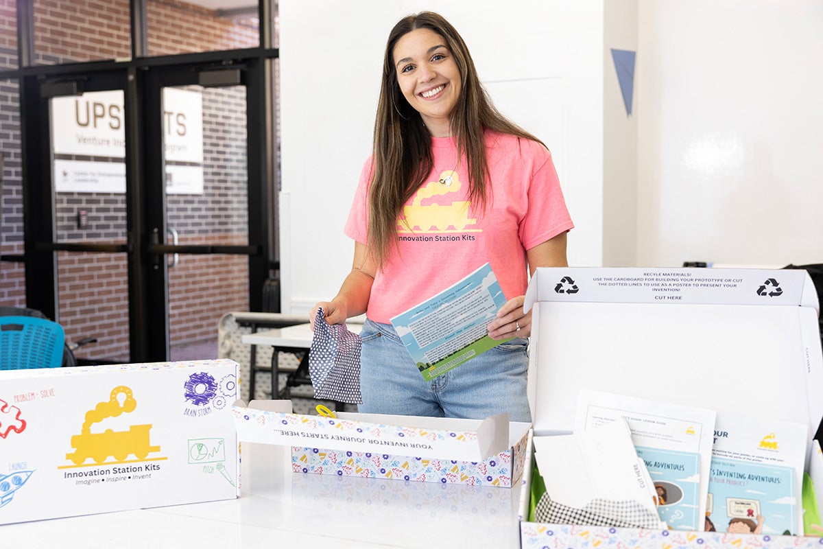 Kelly Shea in pink t shirt stands behind table with innovation kit supplies spread out in front of her