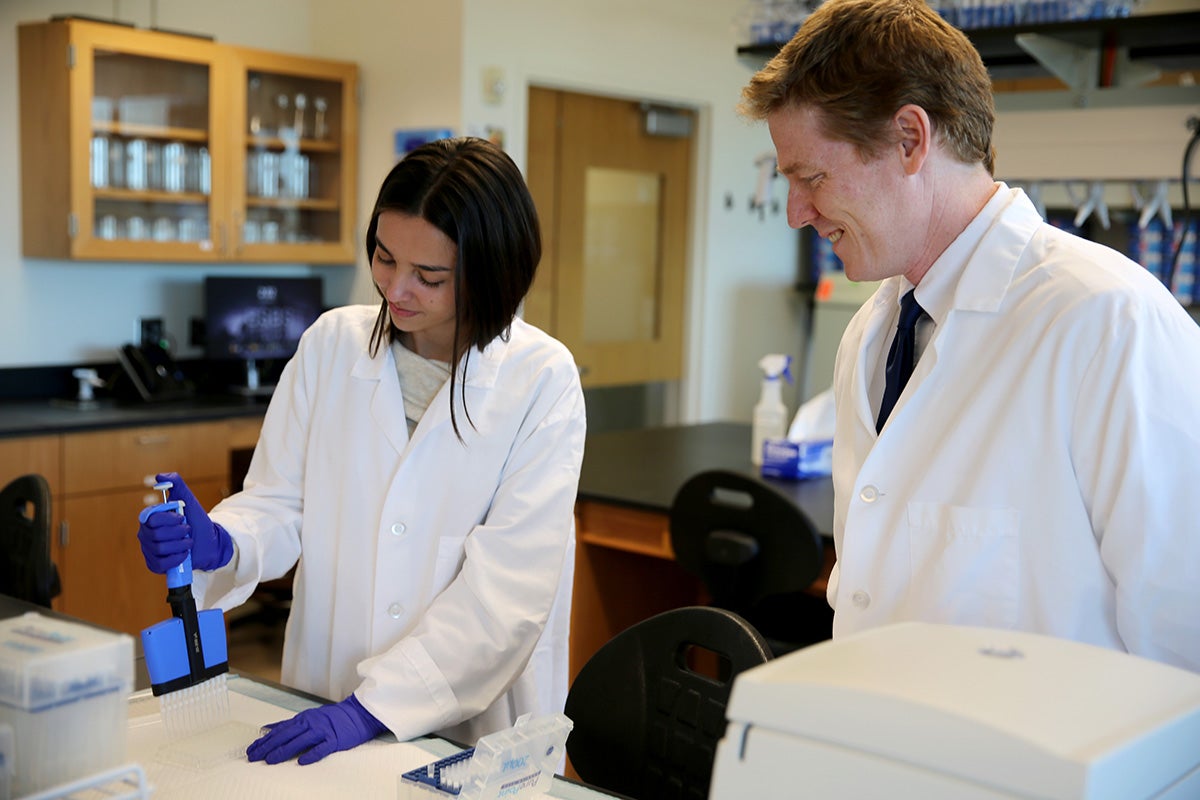 Man in lab coat observes young woman in lab coat, wearing blue gloves and holding medical lab tools, at a table in lab setting