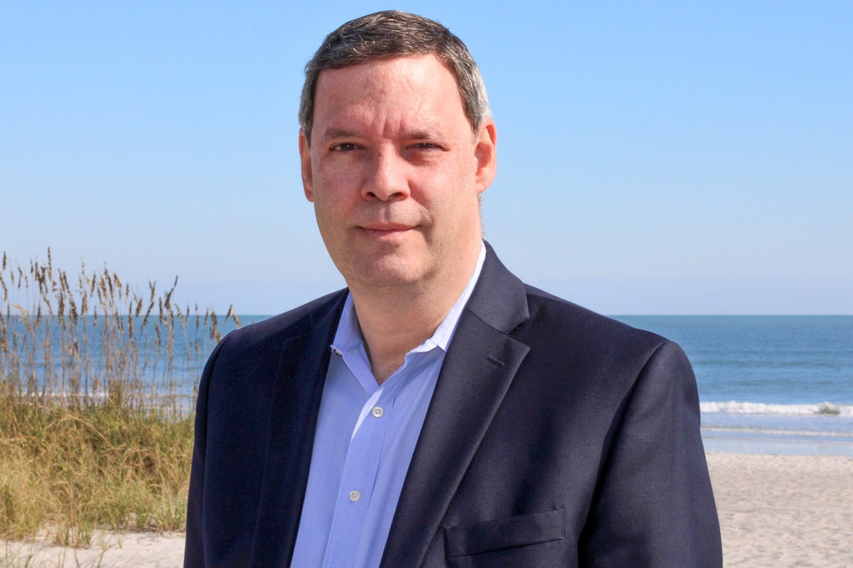 Man in a business suit stands in front of sand dunes and the ocean on a sunny day