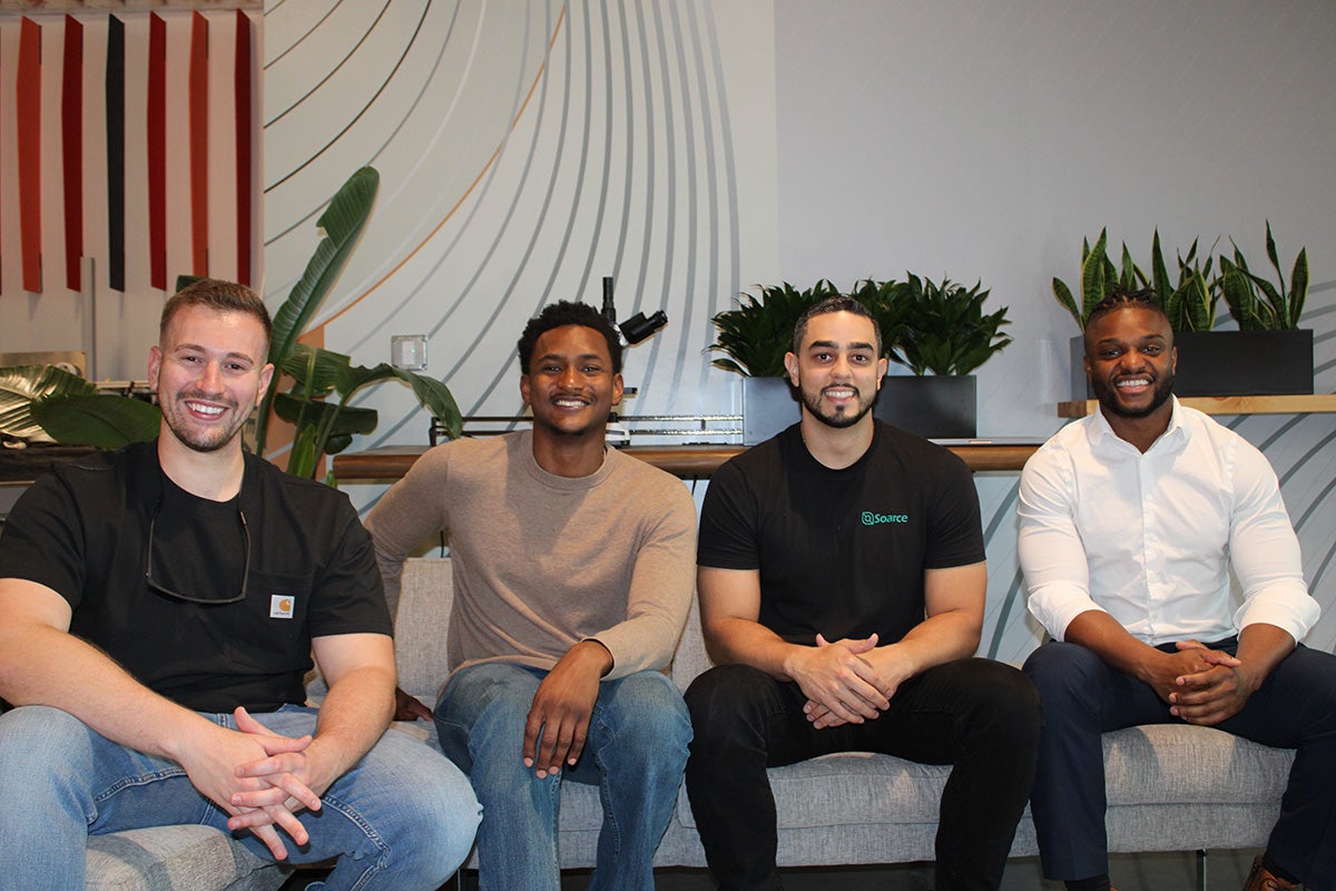 Four men sit on a gray couch side by side with plants on the ledge behind them