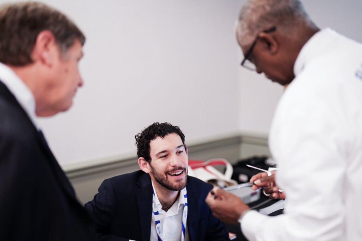Black man in white lab coat and glasses shows metal tool to young bearded man who is sitting