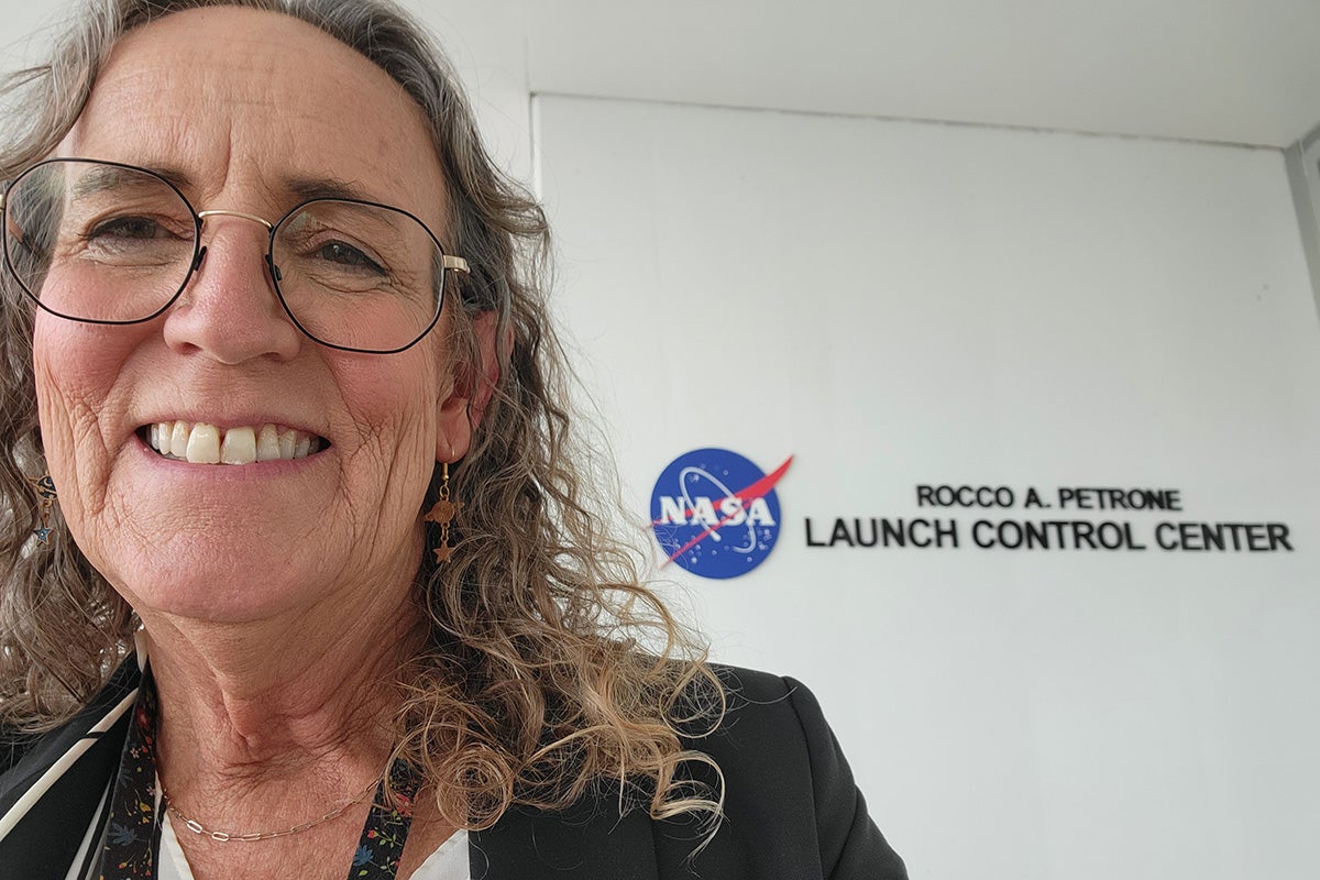 Selfie of woman with curly hair and glasses, standing in front of NASA Launch Control Center signage on white wall