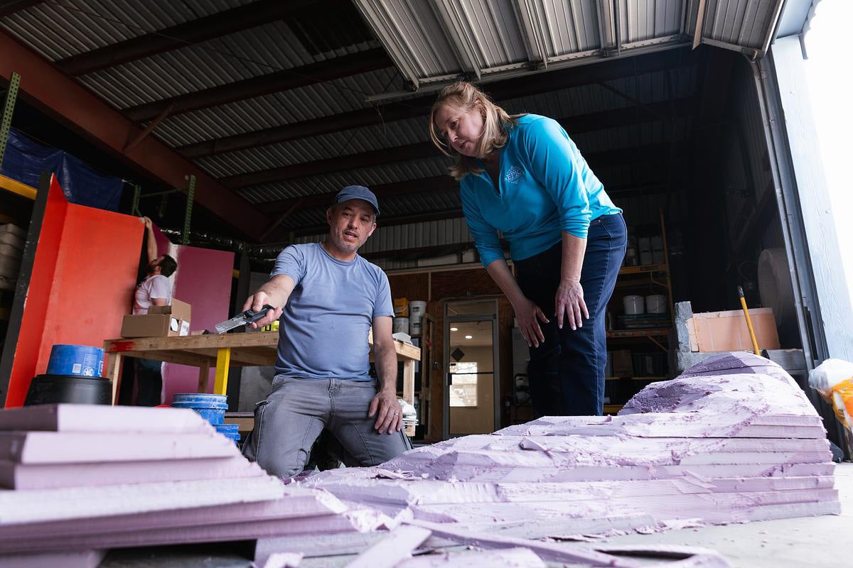 Woman in blue long sleeve shirt stands over a man kneeling with a paint brush in his hand in a workshop