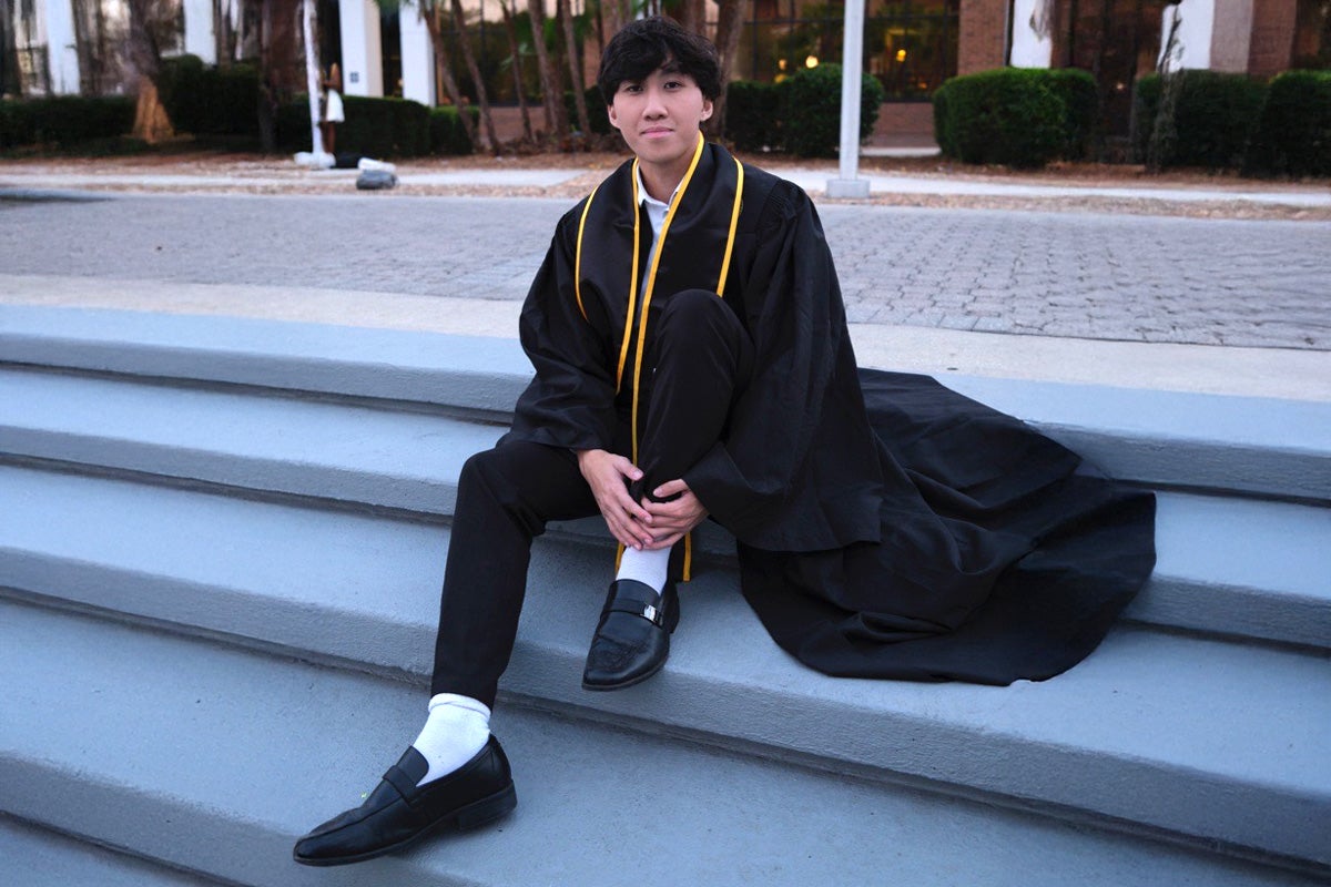 Young man wearing traditional graduation robe sits on concrete steps