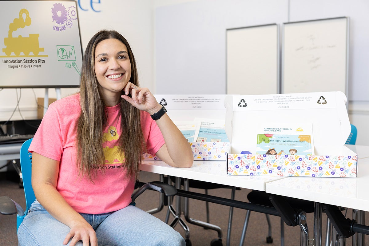 Kelly Shea, in pink t shirt and jeans, sits at a table that displays two innovation kits