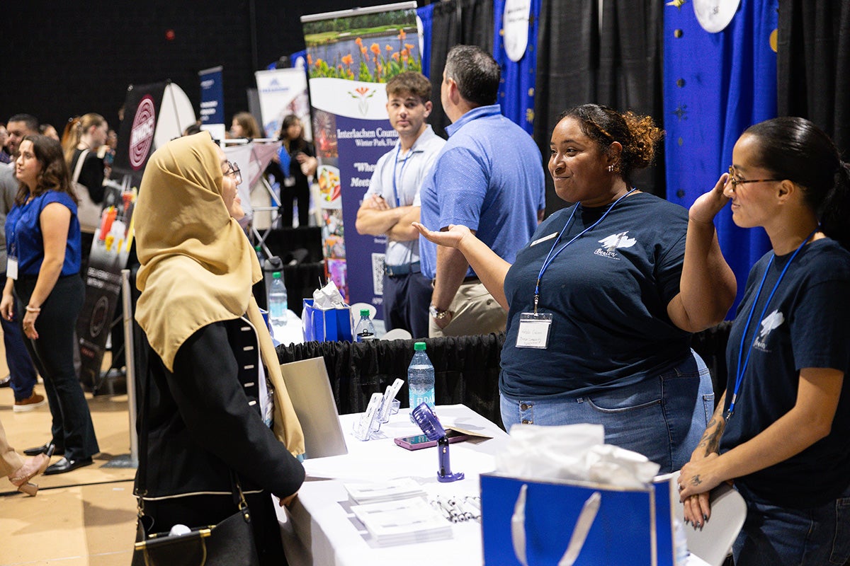 Woman with gold head covering speaks to two officials at a table at career expo in Addition Financial Arena