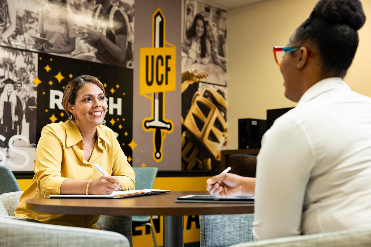 Two women sit at a table across from each other, each holding a pen on a notebook, in front of a UCF-branded wall