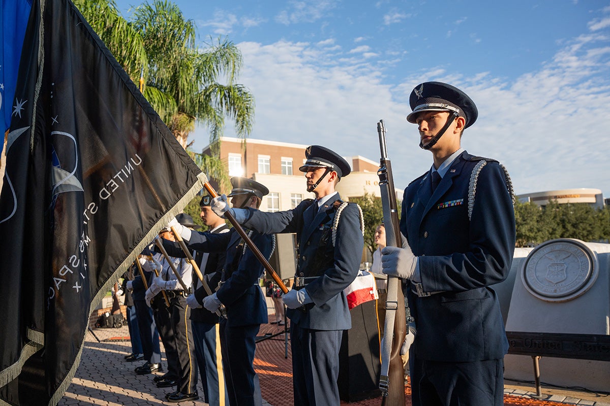 Five military students stand shoulder to shoulder at attention while holding flags on Memory Mall
