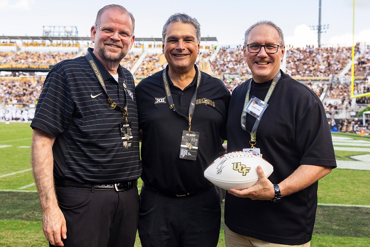 Three men in black UCF polo shirts stand on football field