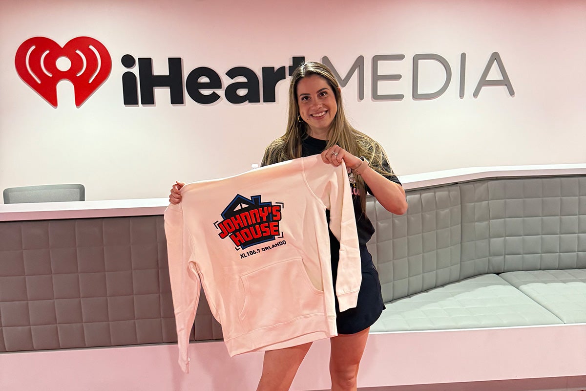 Woman holds up white sweatshirt with Johnny's House logo in front of iHeartMedia sign on white wall