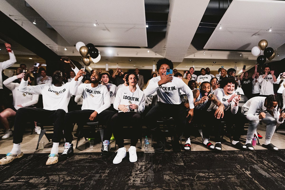 UCF men's basketball team, wearing white long sleeve shirts and black pants, shows joy while seated