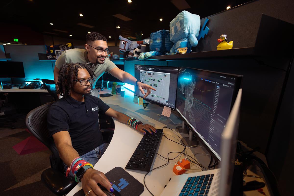 Man wearing glasses stands over another man seated at a desk with three computer monitors