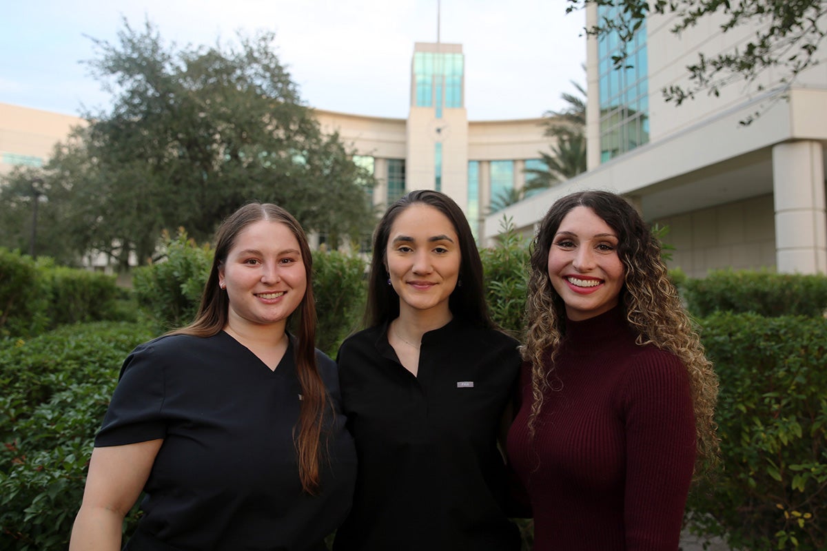 Three brunette women stand side by side in front of green shrubs