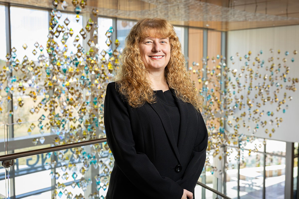Woman with curly-shoulder length hair in black professional suit stands with arms crossed at the waste in front of hanging mosaic art in windowed atrium