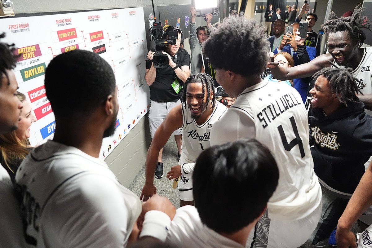 Men's basketball players wearing white UCF jerseys stand by white board bracket ready to put UCF sticker on bracket