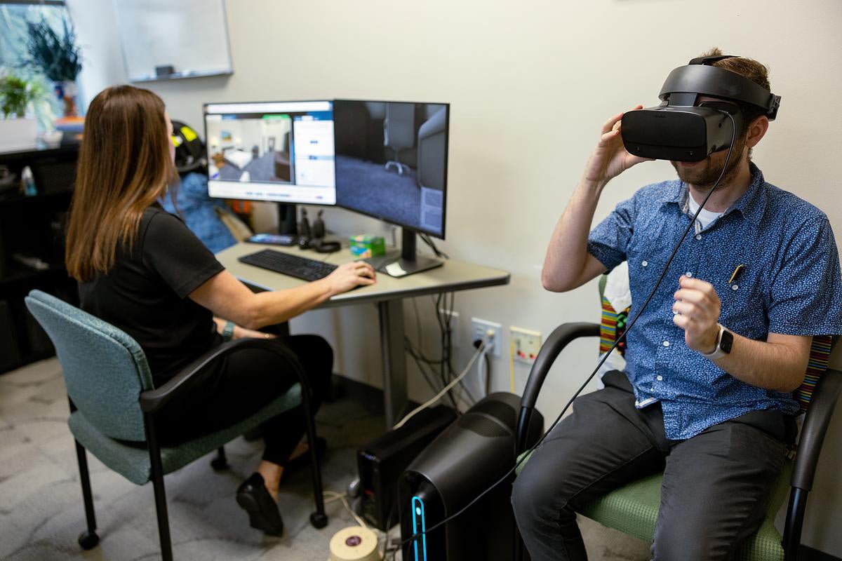 A woman sits at a desk with two computer monitors while a man in blue shirt wearing a black VR headset sits next to the desk.