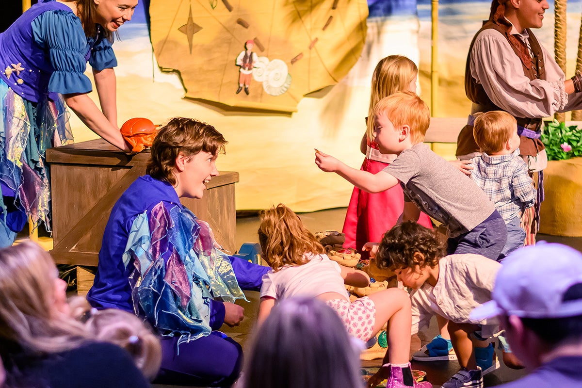 A young blonde boy shows an object in his hand to a performer in a blue and purple costume, surrounded by an audience