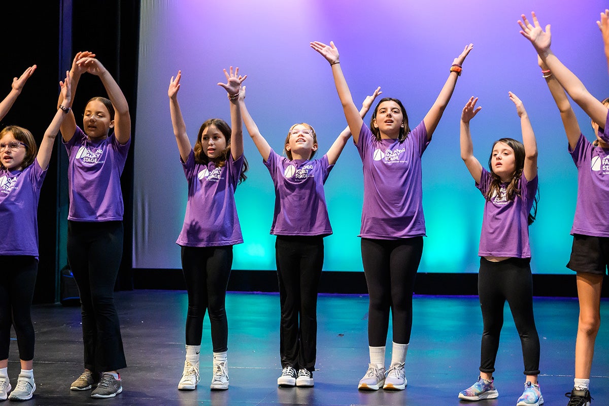 Six girls in purple Orlando Family Stage shirts and black tights stand with arms raised overhead with purple backdrop behind them.