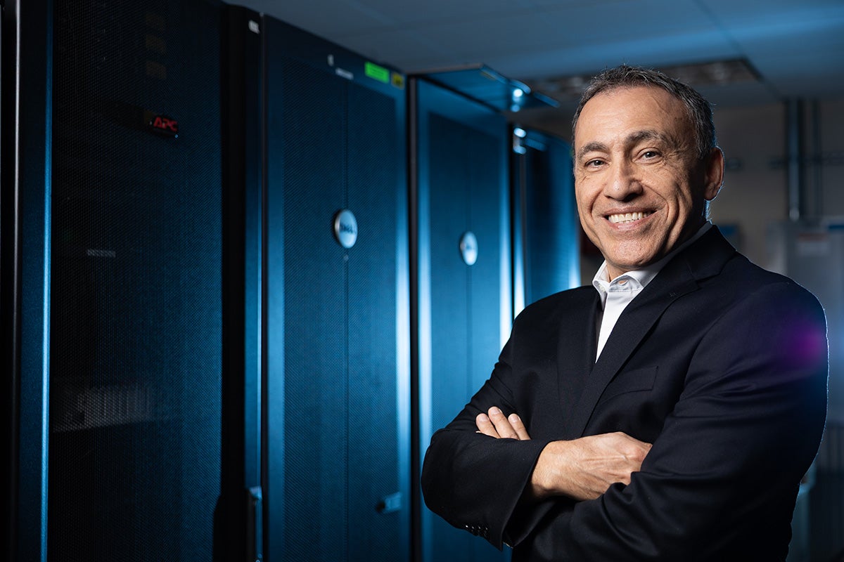 Man in black business suit poses with arms crossed in blue-lit room with Dell server pillars behind him
