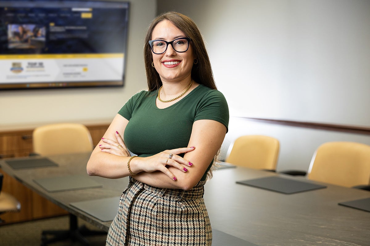 Brunette woman wearing glasses, green shirt and plaid skirt stands in conference room with large table and yellow chairs