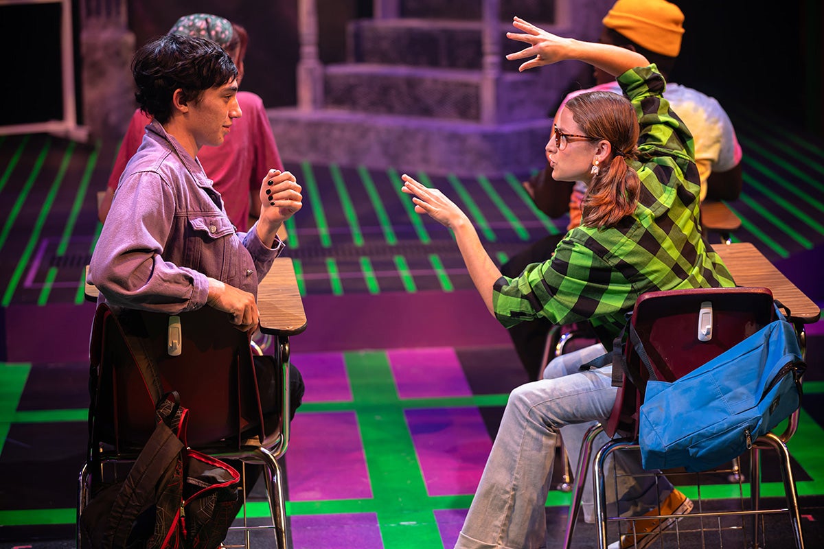 A man and woman sit at two desks across from each other on stage.