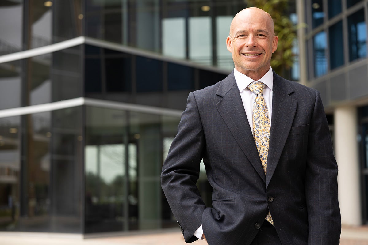 Bald man in business suit and yellow tie stands with right hand in pocket in front of glass and steel building