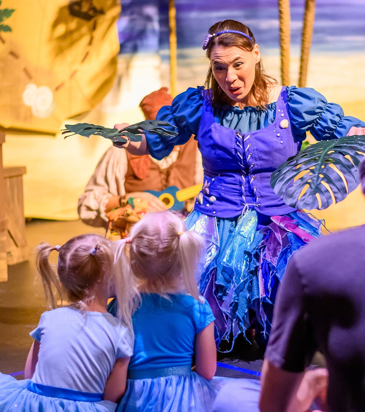 Woman in blue and green costume dress holds palm leaves to two young girls sitting and watching her