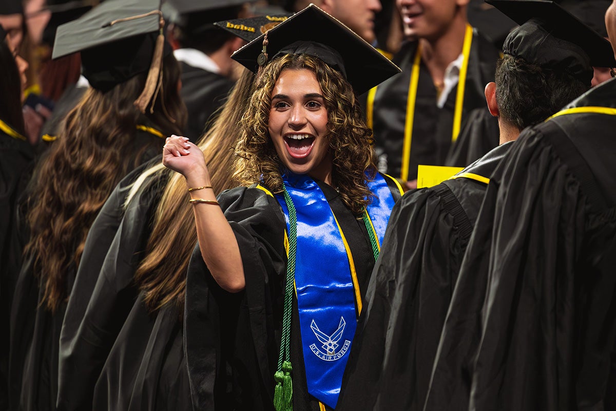 Woman with curly brown hair dressed in black graduation cap and gown with blue decorative Air Force stole smiles in a crowd