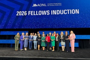 Members of the 2026 Class of Fellows of the American Organization for Nursing Leadership stand together on stage holding awards during the induction ceremony, beneath a large screen displaying “2026 Fellows Induction.”