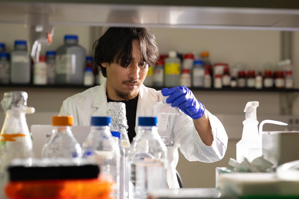 Man with dark hair and wearing a white lab coat and blue latex gloves inspects a glass beaker in a lab setting
