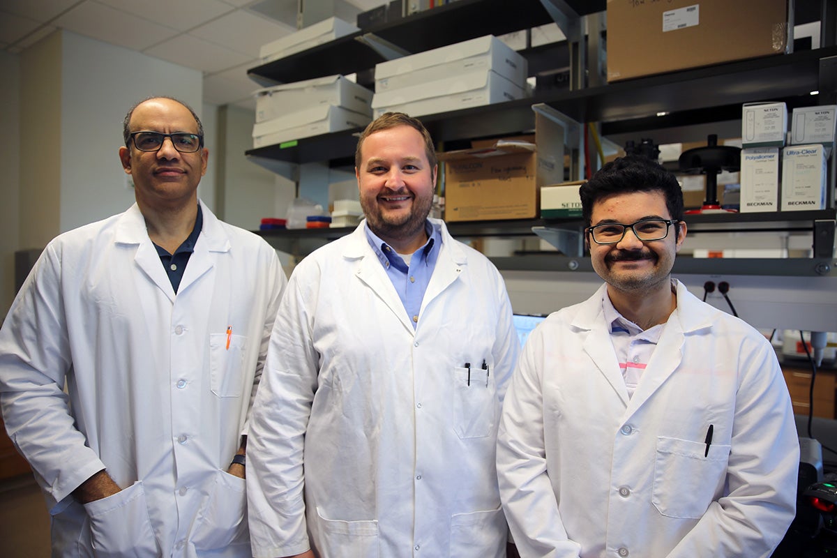Three men stand in white lab coats side by side in front of three rows of shelving with boxes stacked