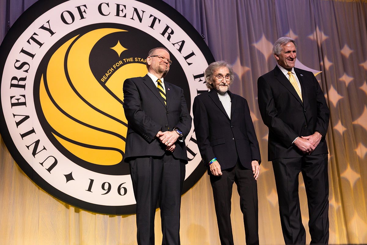 Three men in business suits stand side by side on stage in front of large white/black/gold UCF official Pegasus seal