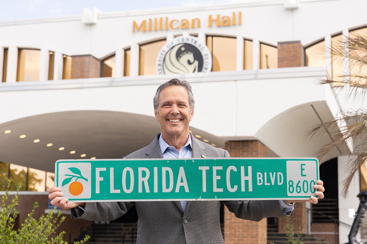 Fred Kittinger in gray suit and blue dress shirt stands in front of Millican Hall holding a Florida Tech blvd green street sign with orange icon