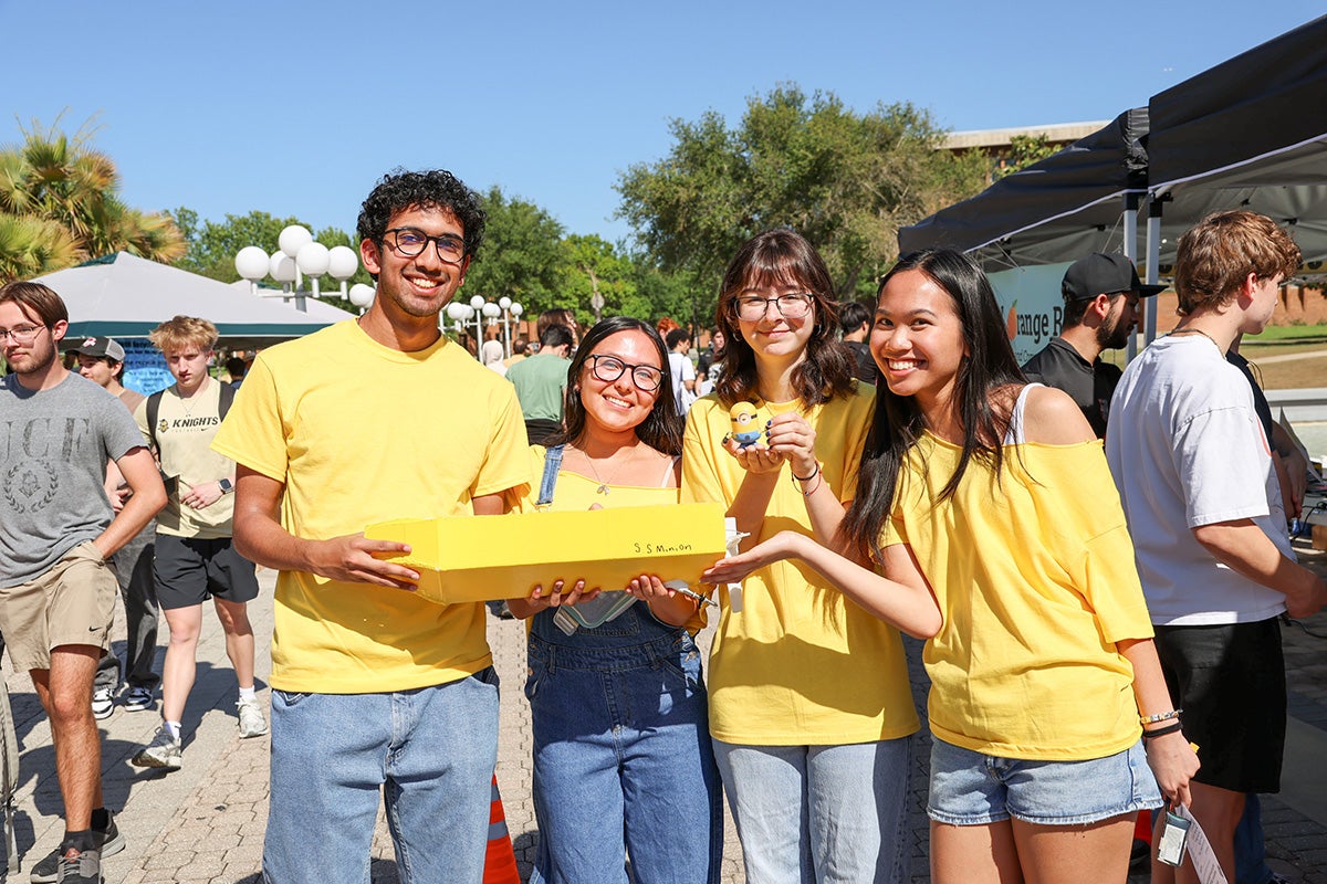 Four college students wearing yellow shirts and jean shorts hold a yellow vessel with "SS Minion" written on body of boat 