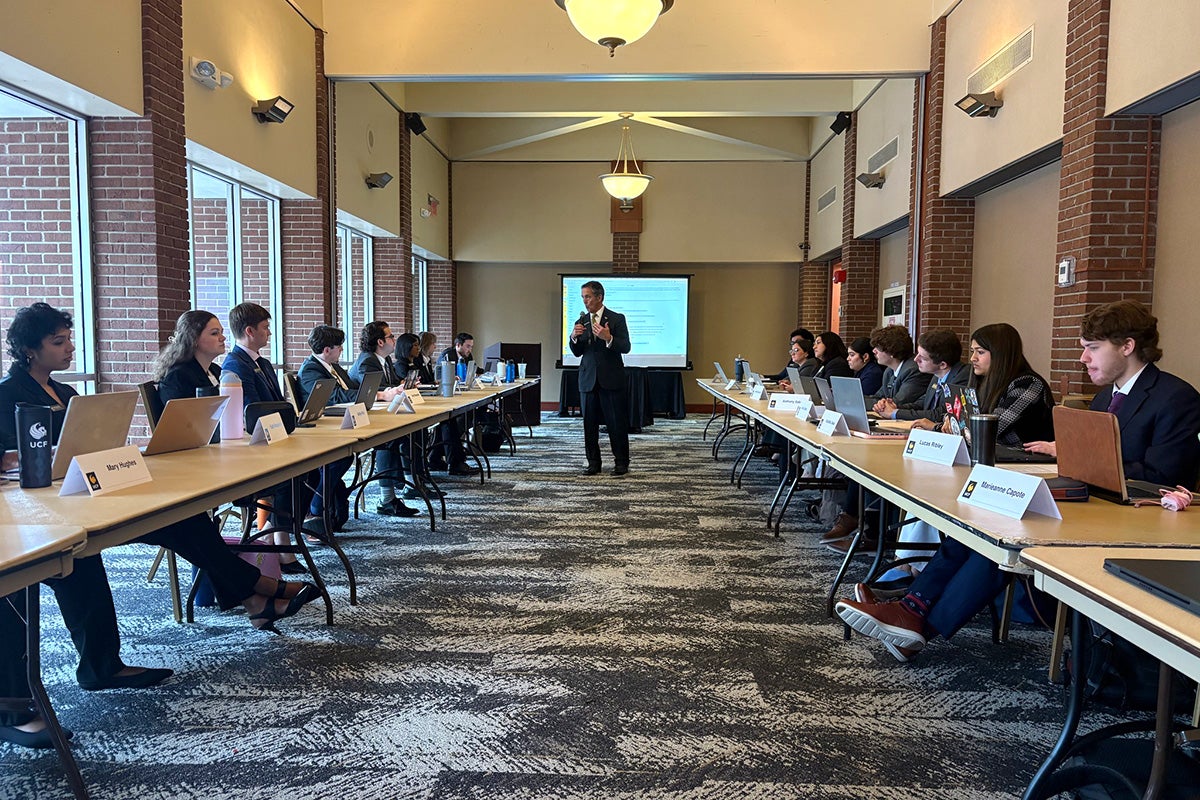 Fred Kittinger stands in between two rows of folding tables with students in business suits seated with namecards in front of them in a large room 