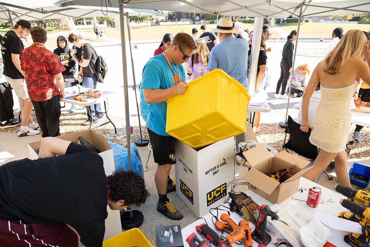 Blonde male wearing bright blue shirt and sunglasses holds yellow bin under a tailgate tent, surrounded by tools and recycled materials.
