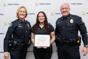 UCF Police Chief Robbin Griffin-Kitzerow and Deputy Chief Scott Freeman stand with a woman as she holds her promotion certificate in front of a UCF Public Safety backdrop.